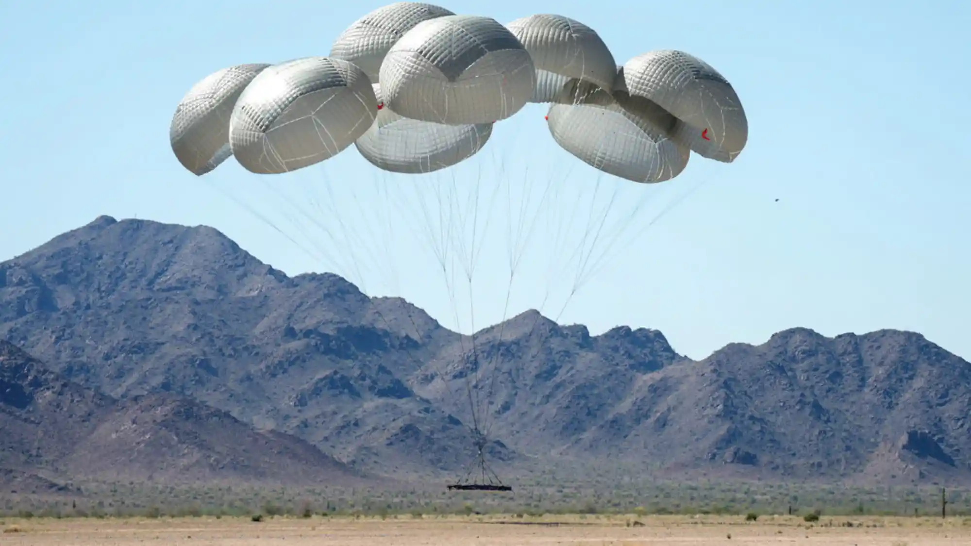 Cluster of several round cargo parachutes over desert and mountains, the load just above the ground