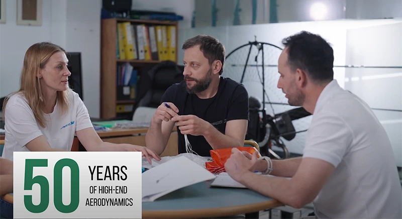 Three people discuss a small orange parachute sample at a table; overlay reads “50 Years of High-End Aerodynamics”