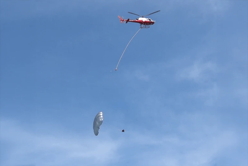 Hubschrauber mit langem Seil am Himmel, darunter ein Fallschirm mit angehängter Last im Sinkflug