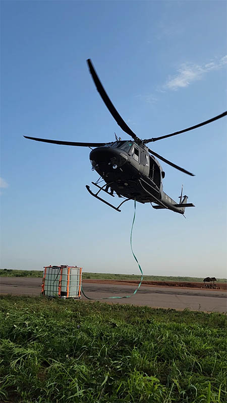 Helicopter hovering over an airfield, a strap running down to a load in a cage container on the ground
