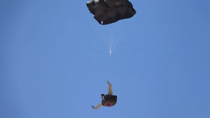 Flying-wing UAV descending under an open recovery parachute against a blue sky