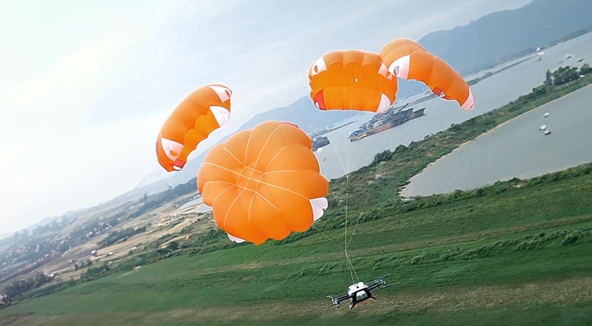 Multicopter drone descending under several orange parachute canopies over a riverside meadow