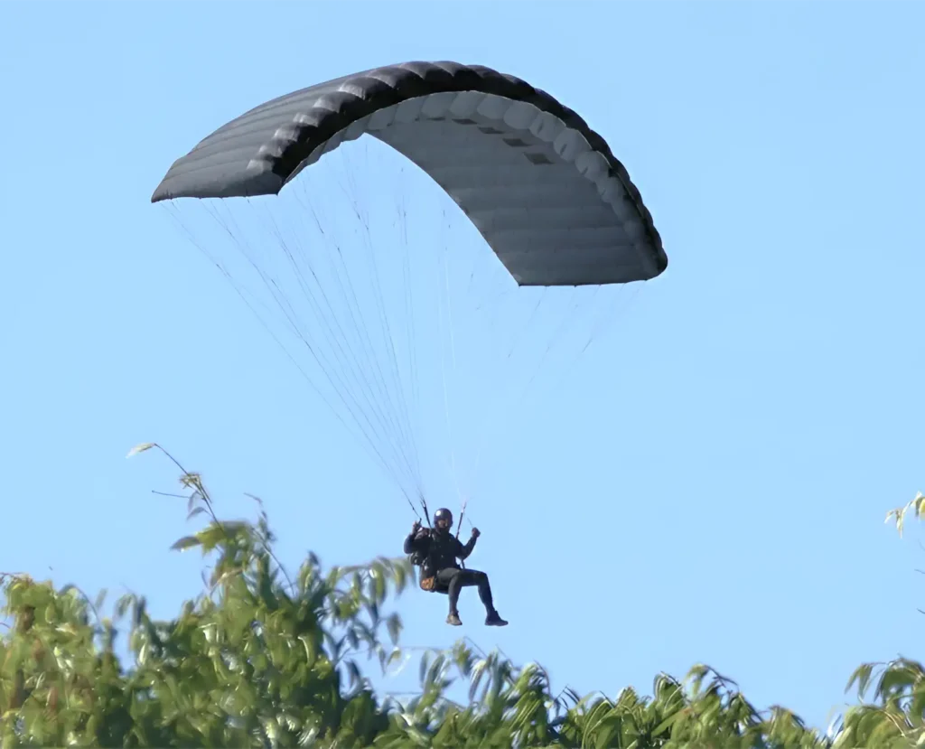 Person under a dark grey ISR ram-air canopy flying over treetops